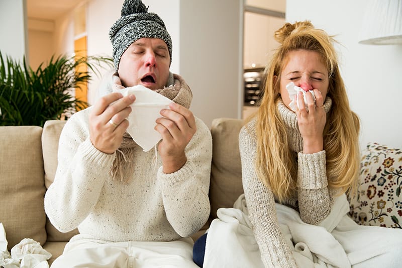 a man and a woman sit on a couch, sneezing into tissues