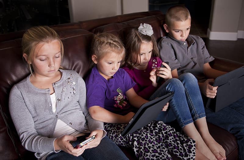 four kids sit on a couch looking at different screens